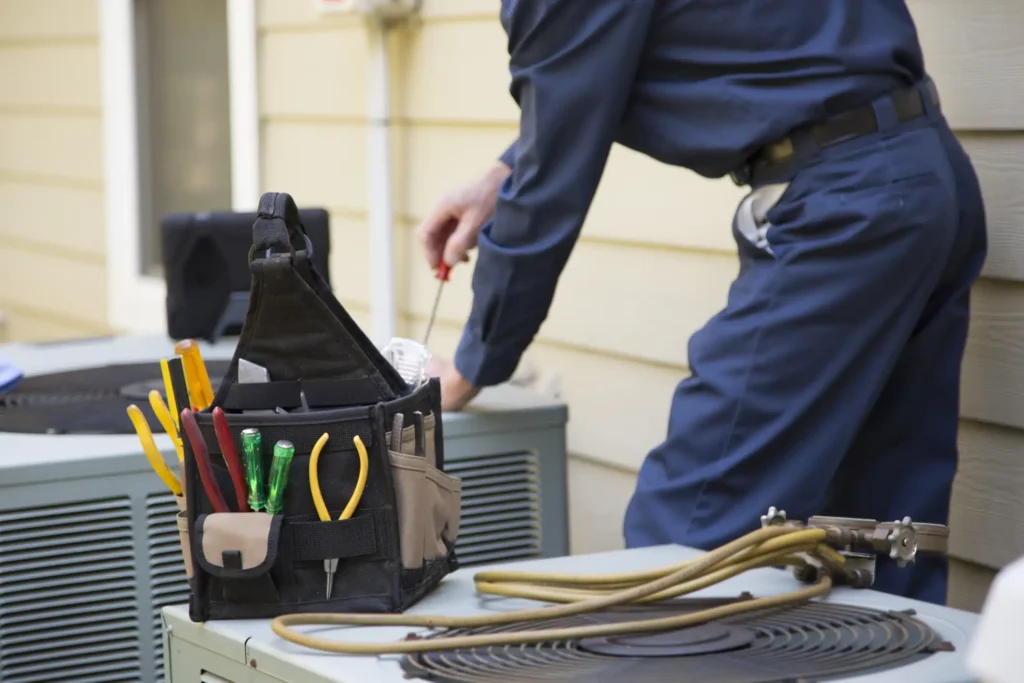 A cropped, close-up shot shows a technician in blue coveralls using a screwdriver to work on an air conditioning unit. In the foreground, a black and tan tool bag sits on top of a different unit, holding various hand tools like pliers and screwdrivers with red, yellow, and green handles. The scene is outdoors, with what appears to be a residential building in the background.