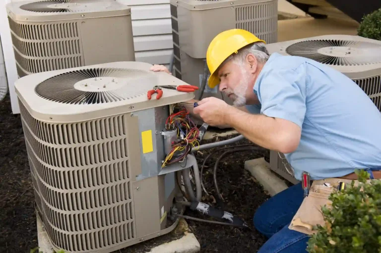 A professional-looking male AC technician, wearing a yellow hard hat, a light blue shirt, and blue jeans, kneels while carefully working on an outdoor air conditioning unit. He holds a screwdriver in one hand, attending to the exposed, colorful wires and components inside the unit. In his other hand, he holds a pair of red-handled pliers, and a tool belt is visible around his waist. Two other similar AC units are visible in the background, suggesting a multi-unit installation or apartment complex.