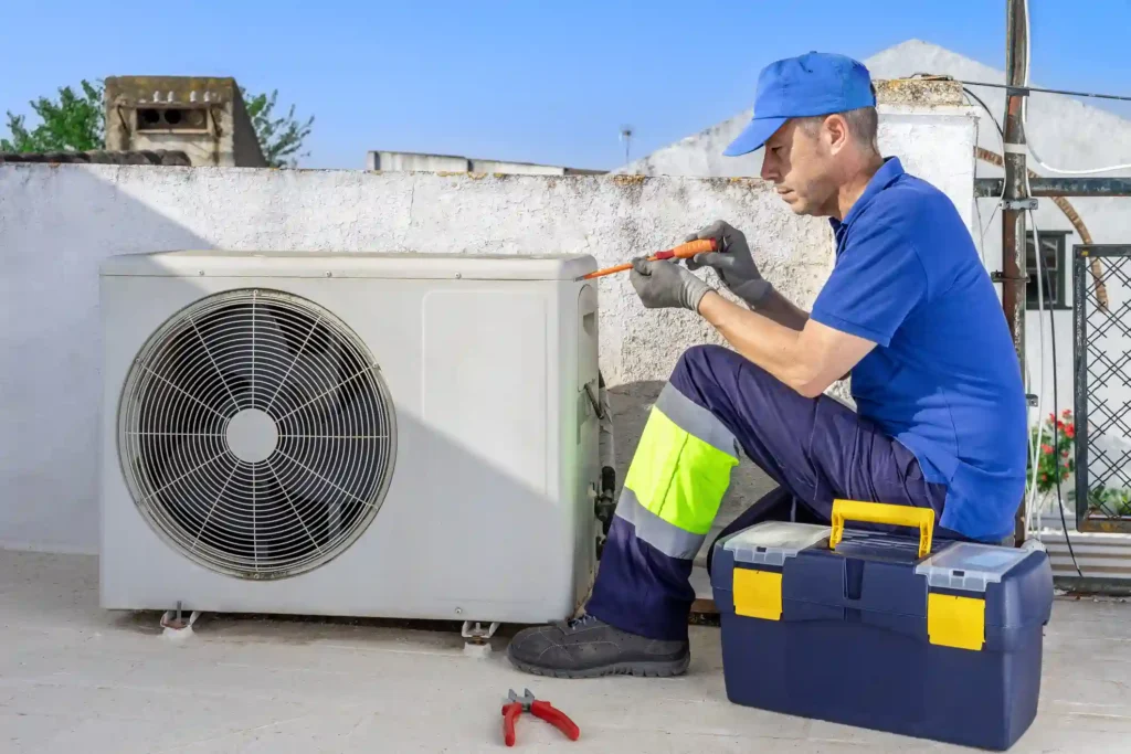 A professional AC repairman, dressed in a blue shirt, blue pants with neon yellow safety stripes, a blue cap, and gloves, kneels on a flat rooftop. He is focused on a large, white air conditioning unit, using a screwdriver to work on the side panel. A red-handled tool and a blue and yellow toolbox are visible on the roof next to him. In the background, there are white buildings and a clear blue sky.
