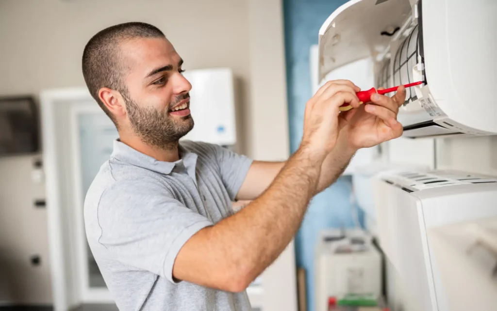 A friendly-looking male AC technician, with a short haircut and a beard, is smiling as he works on a wall-mounted indoor air conditioning unit. He wears a gray polo shirt and uses a red-handled screwdriver to make an adjustment inside the opened casing of the unit. The setting appears to be a residential or commercial interior.
