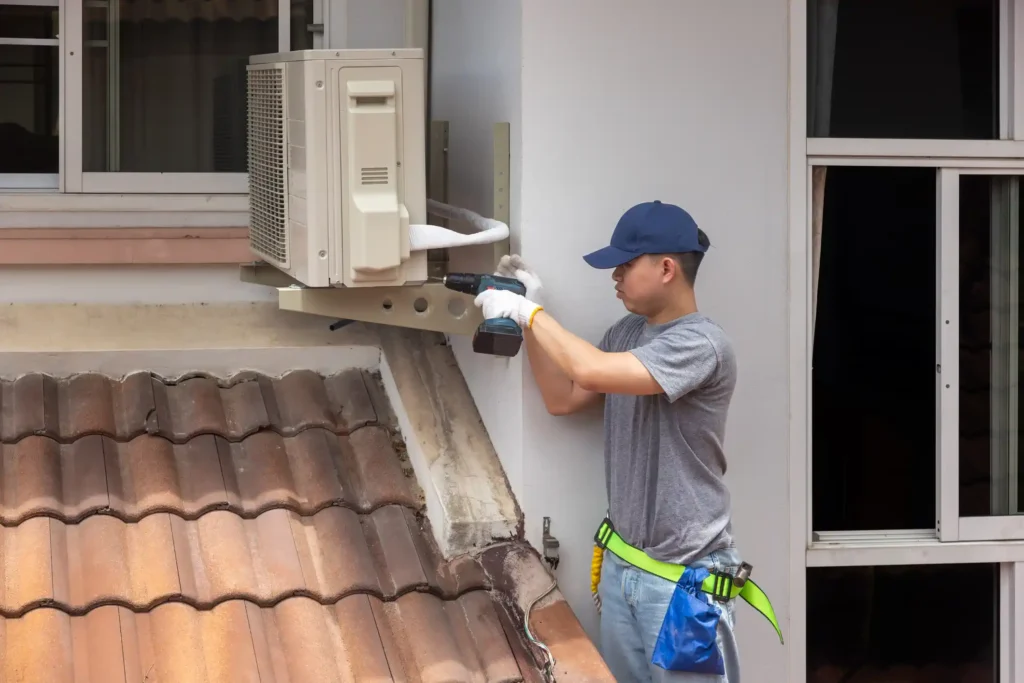 A focused technician, wearing a gray t-shirt, blue jeans, a blue cap, and a tool belt with a bright green strap, is installing or repairing a white AC unit on the exterior of a building. He is using a cordless drill to secure the unit to a wall bracket. The image is taken from a low angle, showing the technician positioned above a clay-tiled roof.
