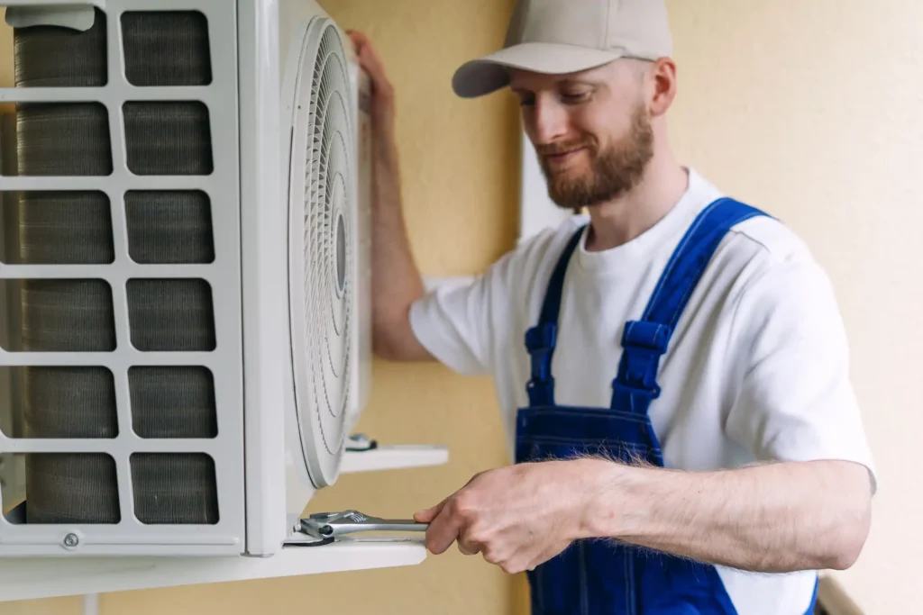 A male technician with a beard and a gray cap smiles while using a wrench to tighten a bolt on a wall-mounted AC unit. He is wearing a white t-shirt under a pair of blue overalls. The photo is a close-up shot that focuses on his hands and the AC unit, which has a large fan visible through a circular grate.