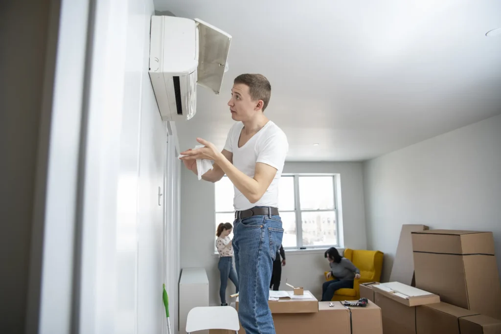 A male technician, wearing a white t-shirt and blue jeans, stands on a small white stool and works on a wall-mounted indoor AC unit. He holds a white cloth or paper in his hands, looking up at the unit with a focused expression. The room appears to be in the process of being moved into or out of, with several cardboard boxes scattered around. In the background, two women are partially visible, one of whom is sitting in a bright yellow chair.