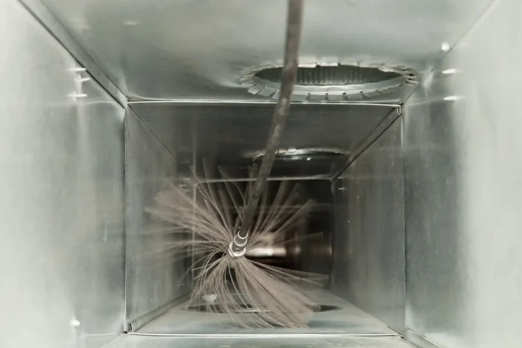 A spinning brush head cleans the inside of a metallic air duct, kicking up dust and debris.
