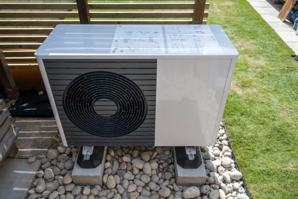 A newly installed outdoor heat pump unit is placed on a bed of pebbles, next to a grassy lawn.
