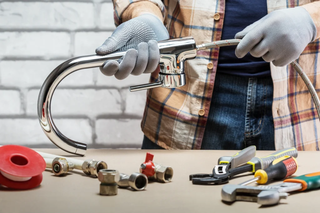 A person in gloves assembling a shiny chrome faucet, surrounded by plumbing tools and parts on a workbench.
