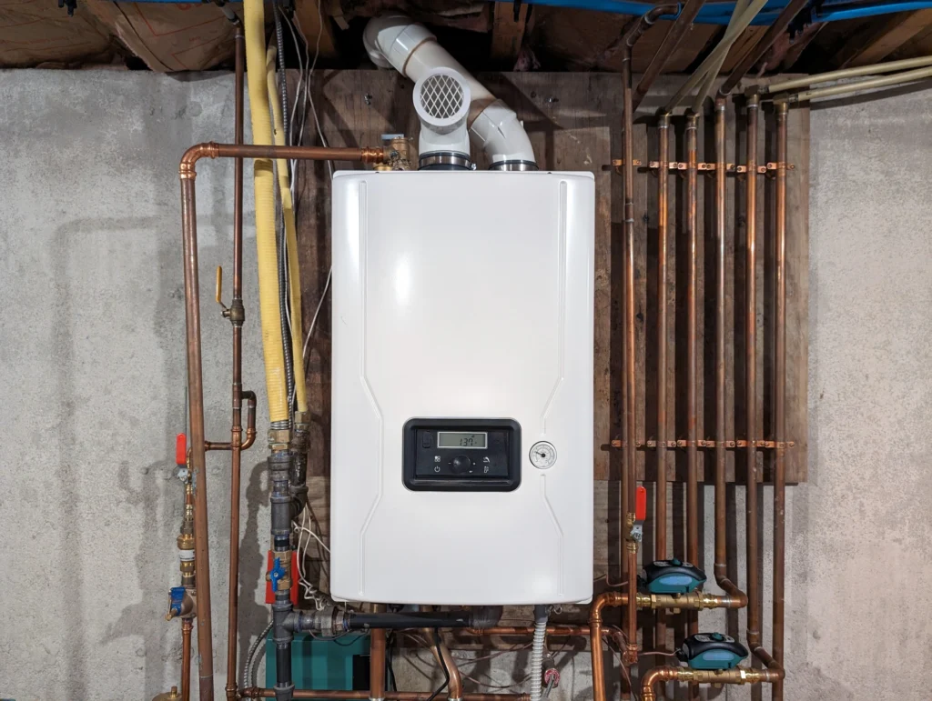 A white tankless water heater is mounted on a basement wall, surrounded by copper pipes and valves.