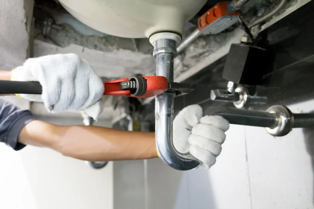 A person wearing white gloves uses a red wrench to tighten a silver pipe under a sink