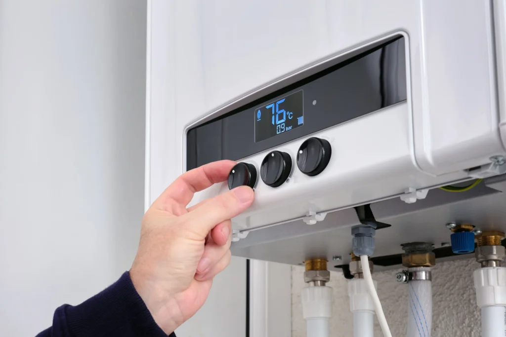 A hand adjusts the temperature knob on a digital water heater display showing 76 degrees Celsius.