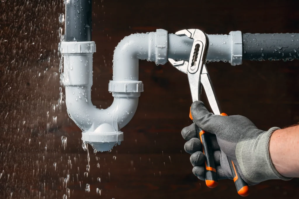 gloved hand grips pliers, fixing a leaking pipe with water spraying out.
